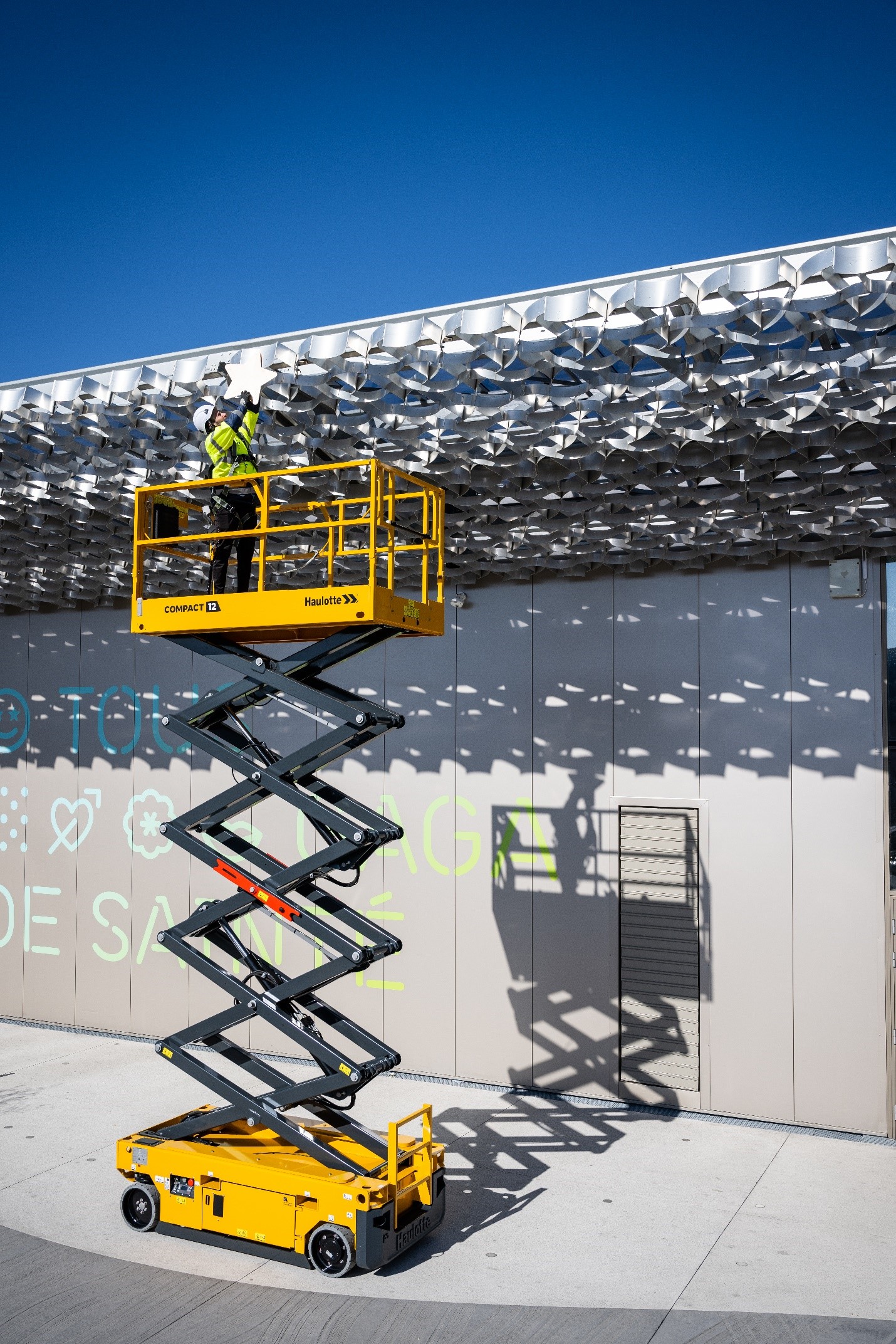 Podnośnik nożycowy Haulotte Compact 12 z pracownikiem wykonującym prace montażowe na elewacji budynku