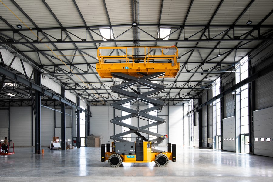 Duży podnośnik nożycowy Haulotte HS18 E PRO, uniesiony na maksymalną wysokość wewnątrz hali przemysłowej